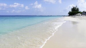 Ein Strand mit klarem türkisblauem Wasser, weißem Sand und einigen Bäumen sowie Gebäuden im Hintergrund.