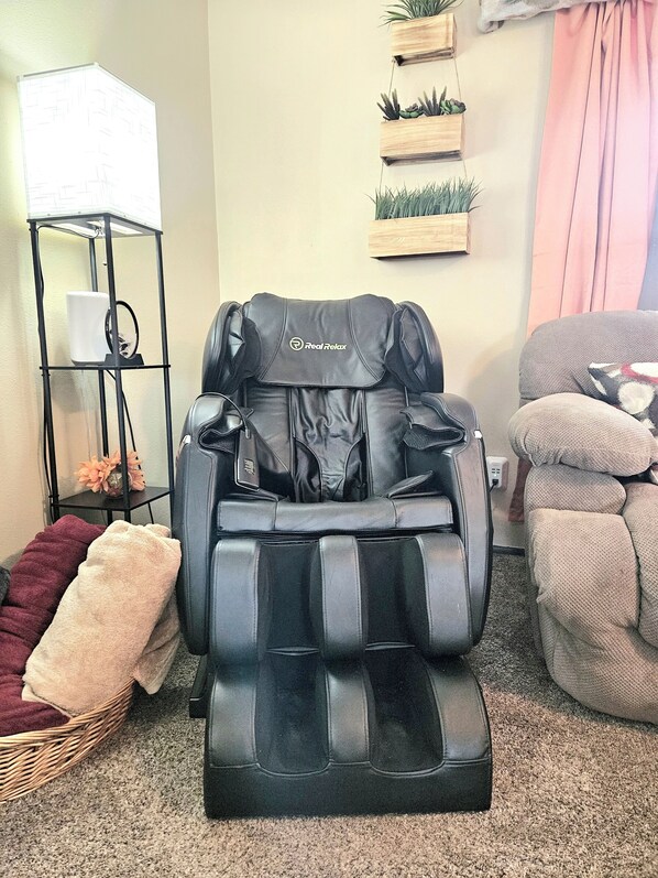 A massage chair with a control panel, a small shelf with decorative items, and a wicker basket with a towel. - Spacious Family Getaway • Hot Tub • Parks & Trails (Arvada)