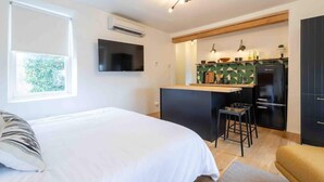 A bedroom with a large bed, a TV mounted on the wall, and a kitchen area with a refrigerator, bar stools, and a countertop.