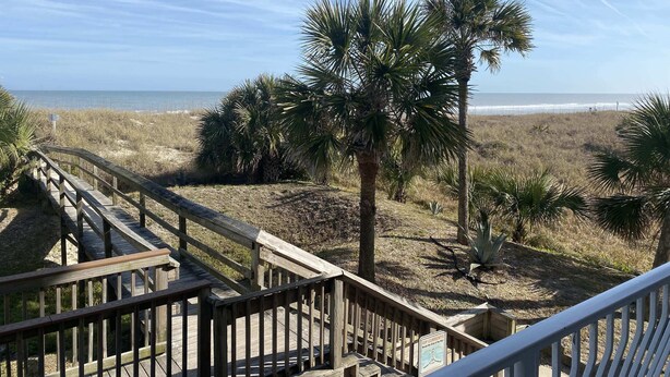 Eine Holztreppe führt zu einem Strandabschnitt mit Palmen und Blick auf das Meer.