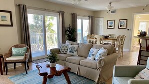 Ein Wohnzimmer mit beiger Couch, einem Holztisch und einem Streifen Teppich. Es gibt eine Essecke mit Tisch und Stühlen sowie einen Balkon mit Vorhängen.