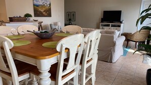 Ein Essbereich mit einem Holztisch und weißen Stühlen, einem Fernseher im Hintergrund und einer Pflanze rechts.