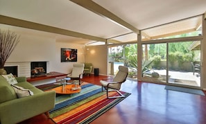 A modern living room with a fireplace, a colorful striped rug, and a view of a backyard with a pool.