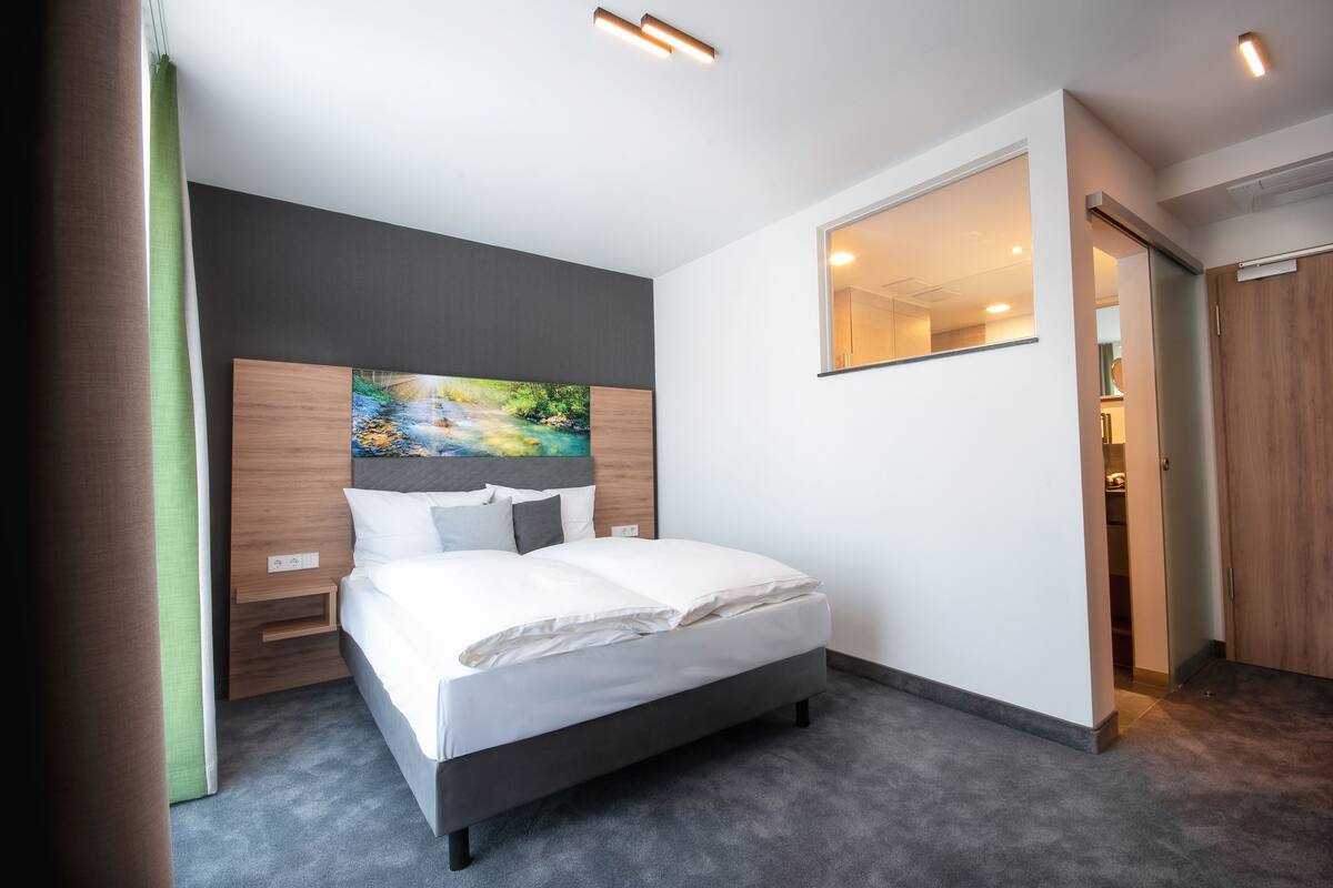 A modern hotel room with a large bed, a painting on the headboard, a mirror on the wall, and a wooden door.