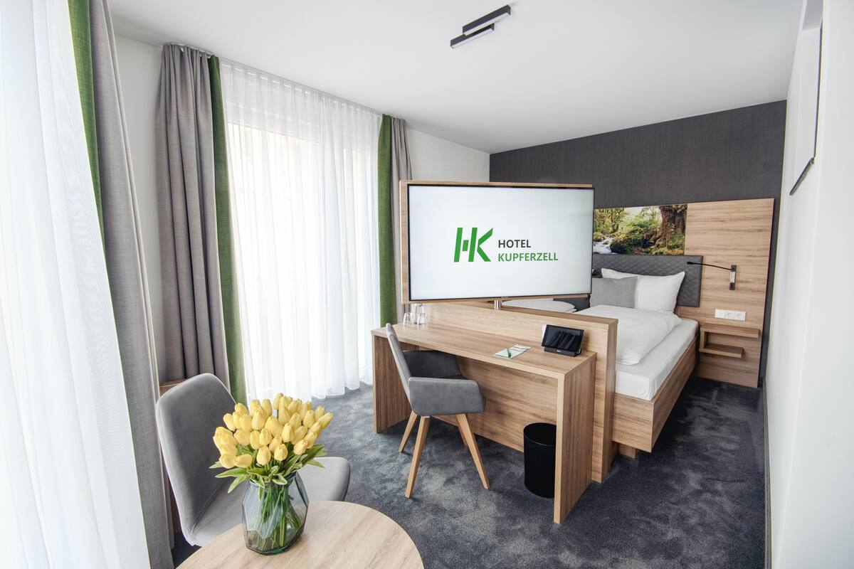 A hotel room with a bed, a desk, a chair, a TV, and a vase of yellow flowers.
