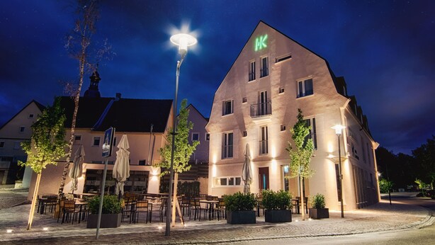 Ein Hotel mit einem auffälligen 'HK'-Schild, einem Sitzbereich im Freien mit Sonnenschirmen und einer gut beleuchteten Fassade.