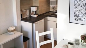 Une cuisine compacte avec des meubles blancs, un four à micro-ondes, une machine à café et un évier. On y trouve une chaise blanche et une petite table avec une tasse et un vase.