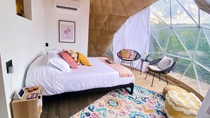 Un dormitorio con cama, almohadas, una mesita de noche con teléfono y cargador, una silla y vistas a través de un ventanal.