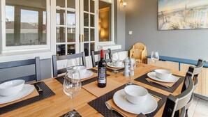 Un comedor con una mesa preparada para cuatro personas, una botella de vino y un cuadro de una playa en la pared.