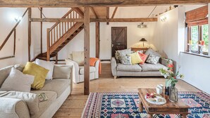 A cozy living room with a wooden staircase, a sofa set, a coffee table with fruits and a vase, and a patterned rug.