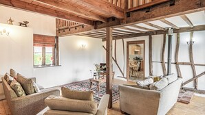 A rustic interior with exposed wooden beams, a cozy seating area with sofas and a patterned rug, and a view into a kitchen through an open doorway.