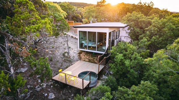 Una moderna cabina con una camera da letto interamente in vetro, un terrazza in legno e una vasca idromassaggio, immersa in una fitta foresta.