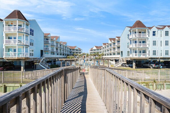 A boardwalk leading to a row of multi-story buildings with balconies, a wooden walkway, and a clear sky.