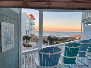 A balcony with teal chairs, a sign reading "Sunrise, Sunburn, Sunset Repeat," and a view of the ocean and a pier.