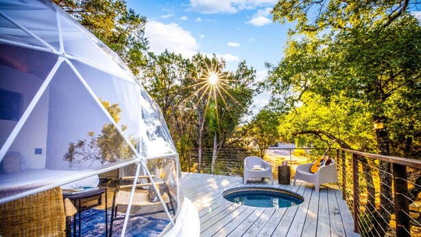 Eine transparente Kuppelkonstruktion mit Pool und Sitzgelegenheiten auf einer Holzterrasse, umgeben von Bäumen.