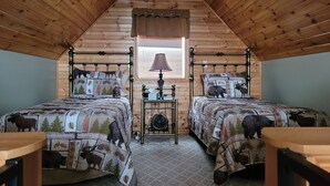 A cozy attic bedroom with wooden walls and a sloped ceiling. It features two beds with animal-themed bedding, a small table with a lamp, and a window with blinds. - Luxury Cabin #3 (Mars Hill)