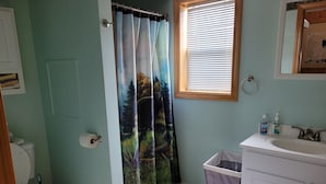 A bathroom with a toilet, a sink, a trash bin, a window with blinds, and a shower curtain with a forest scene. - Luxury Cabin #3 (Mars Hill)