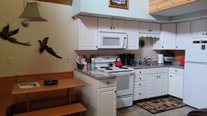 A kitchen with white cabinets, a microwave, stove, and a red kettle. There is a wooden table with a bench and a framed picture on the wall. - Luxury Cabin #3 (Mars Hill)