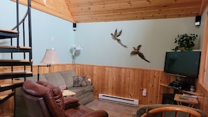 A living room with a brown leather chair, a grey sofa, a wooden TV stand, and a wall-mounted TV. - Luxury Cabin #3 (Mars Hill)
