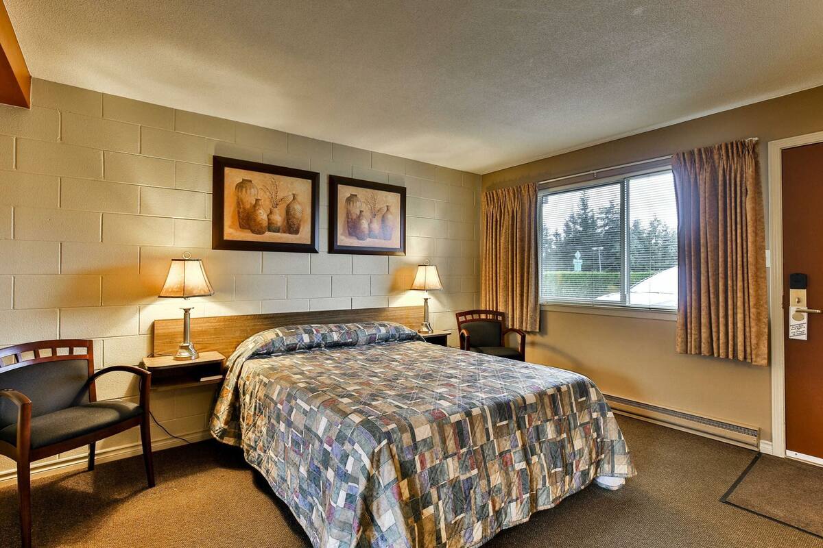 A hotel room with a bed, two chairs, a nightstand, a lamp, a window with curtains, and two framed pictures on the wall.