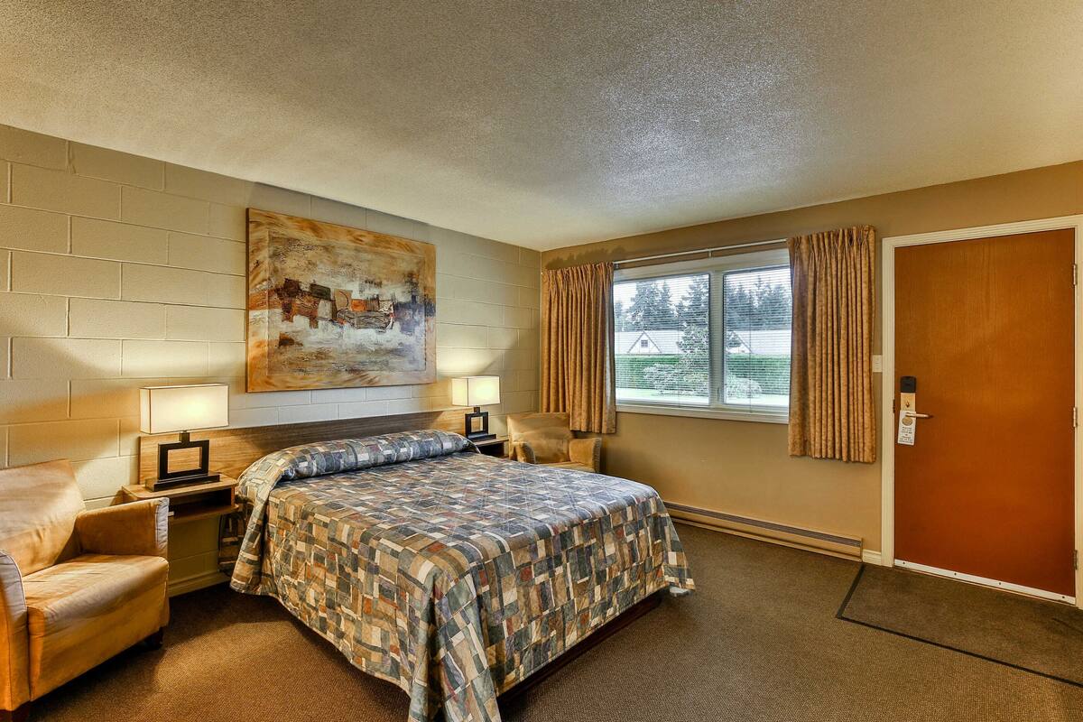 A hotel room with a bed, a chair, a painting on the wall, a window with curtains, and a door.