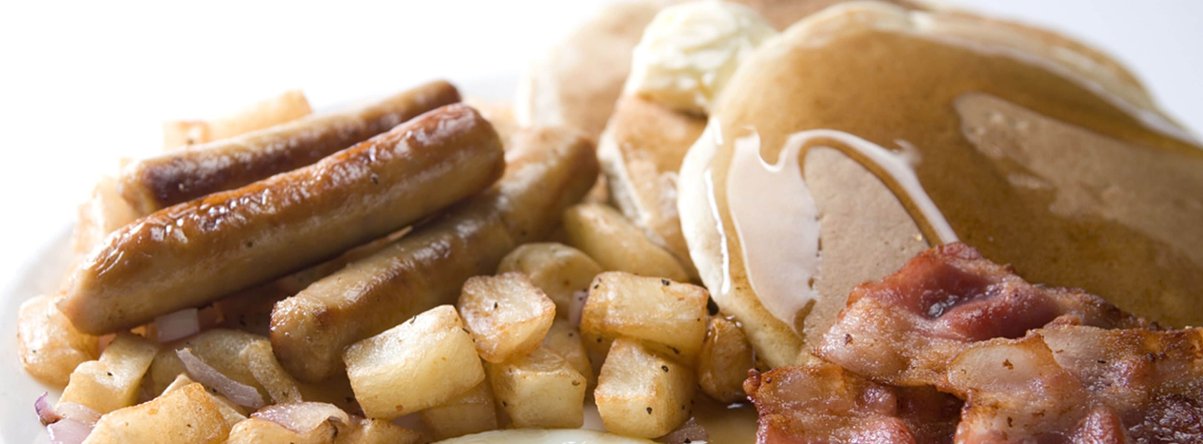 A breakfast plate with sausages, bacon, potatoes, and pancakes with syrup.
