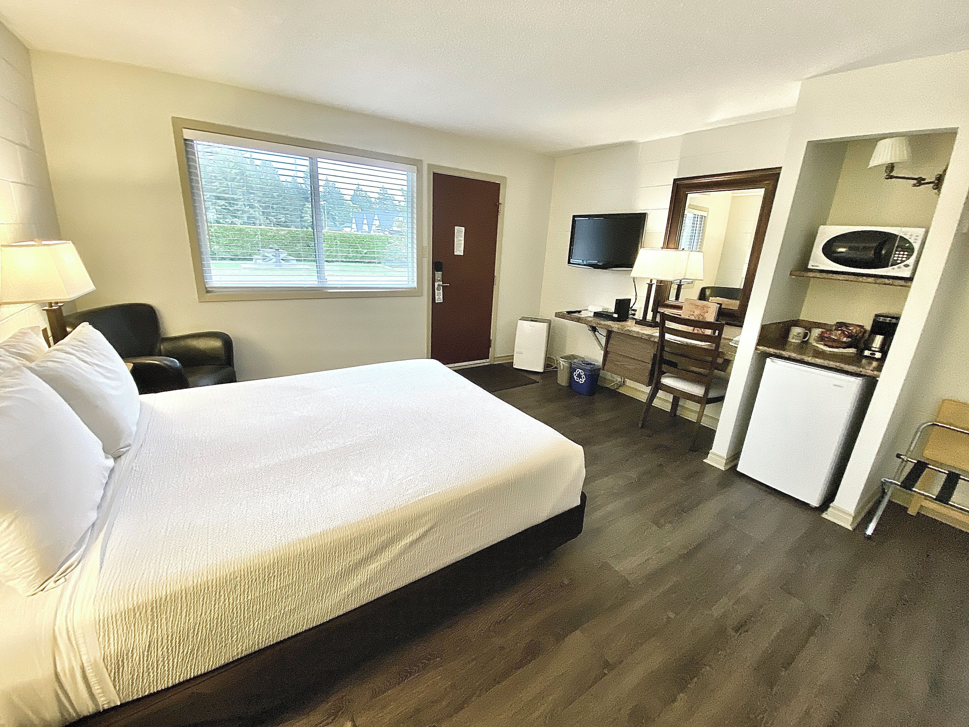 A hotel room with a bed, desk, kitchenette, TV, and a window with blinds.