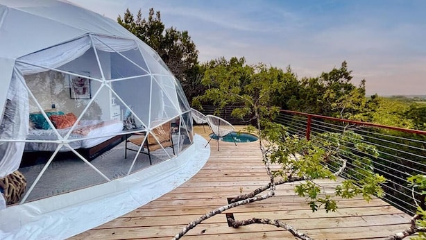 A geodesic dome structure with a bed and seating area, connected to a wooden deck with a pool and surrounded by trees.