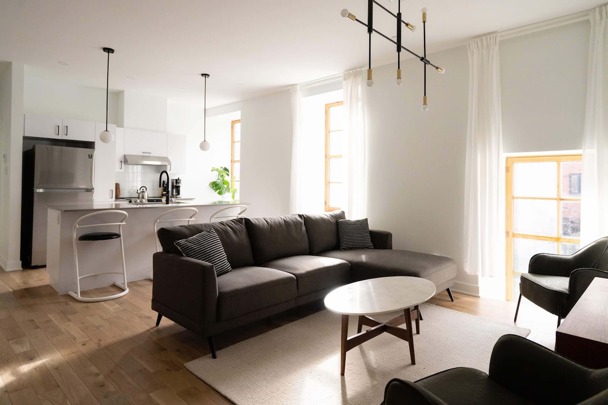 A modern living room with a dark grey sofa, a round coffee table, and a white rug. There is a kitchen area with a stainless steel refrigerator and a white countertop.