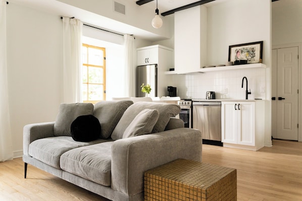 A modern living room with a grey sofa, a small wooden side table, and a kitchen area with white cabinets and appliances. - Maison Rasco by Luxury In Transit (Montreal)