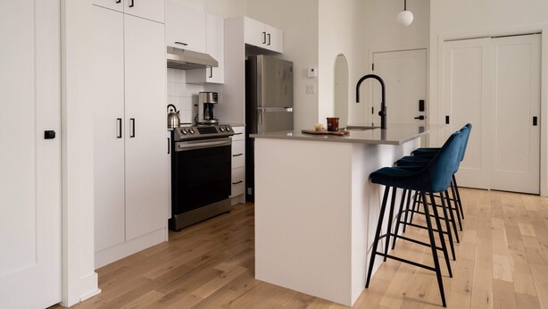 A modern kitchen with white cabinetry, a stainless steel refrigerator, a gas stove, and a coffee maker. There are bar stools and a pendant light above the island. - Maison Rasco by Luxury In Transit (Montreal)