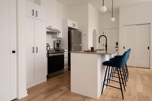 A modern kitchen with white cabinetry, a stainless steel refrigerator, a gas stove, and a coffee maker. There are bar stools and a pendant light above the island. - Maison Rasco by Luxury In Transit (Montreal)