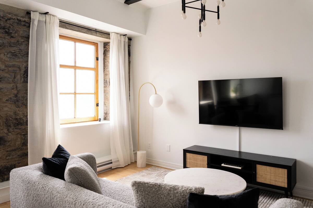 A modern living room with a stone wall, a large window with white curtains, a round white table, a grey sofa, a black TV stand, and a floor lamp.