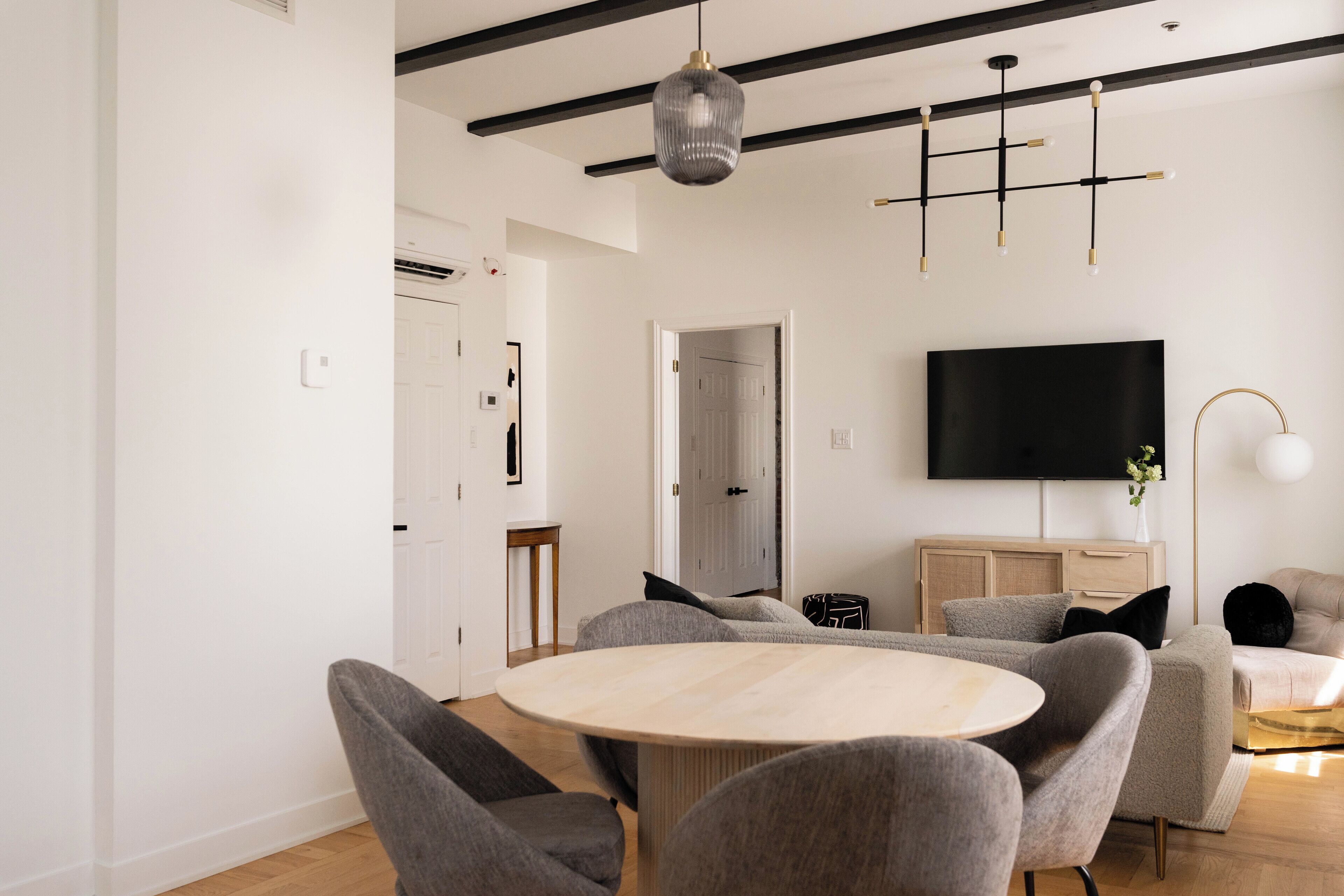 A modern living room with a round wooden table, grey upholstered chairs, a flat-screen TV mounted on the wall, and a minimalist design with exposed beams and a pendant light.