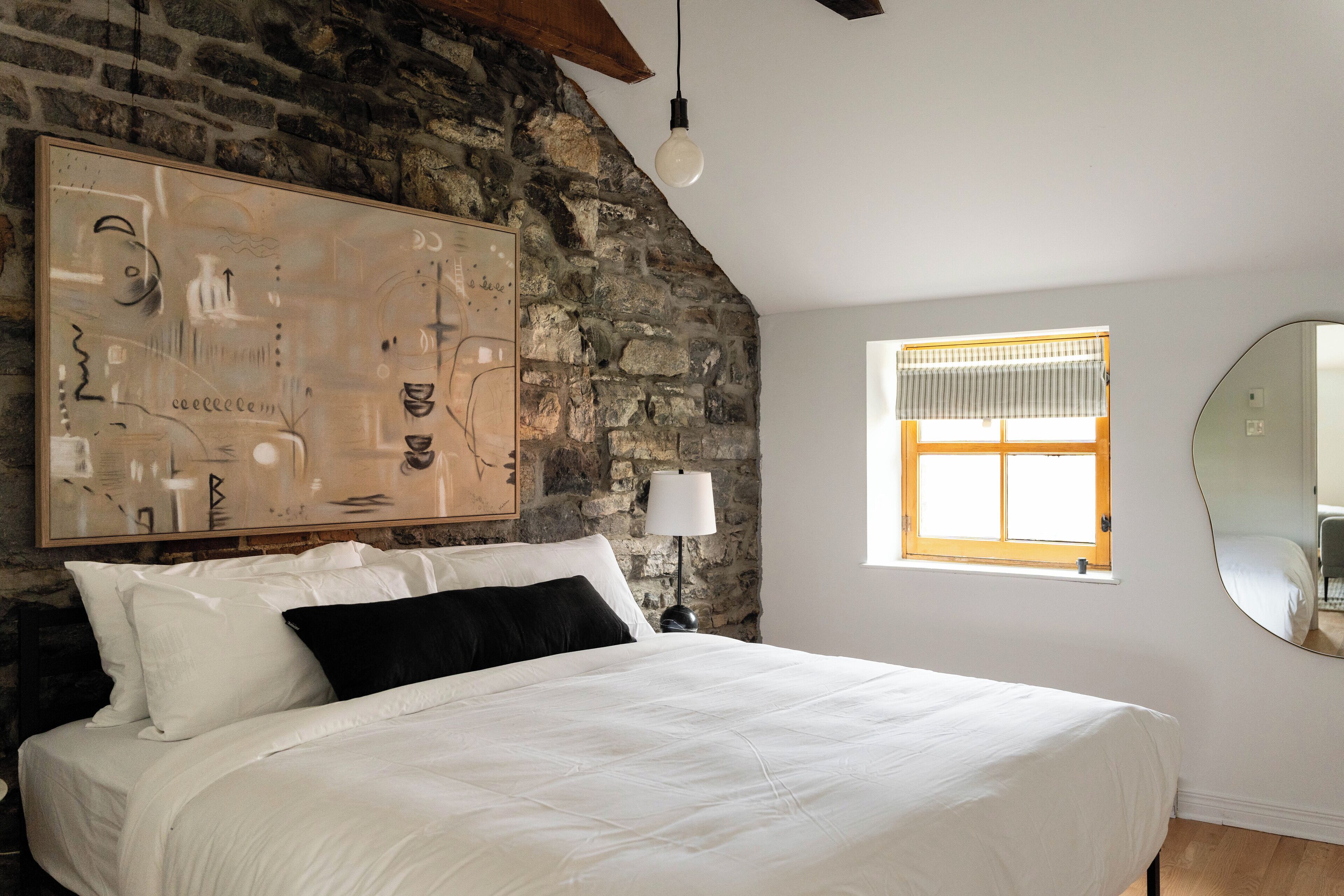 Een slaapkamer met een stenen muur, een groot bed, een raam met jaloezieën en een ronde spiegel.