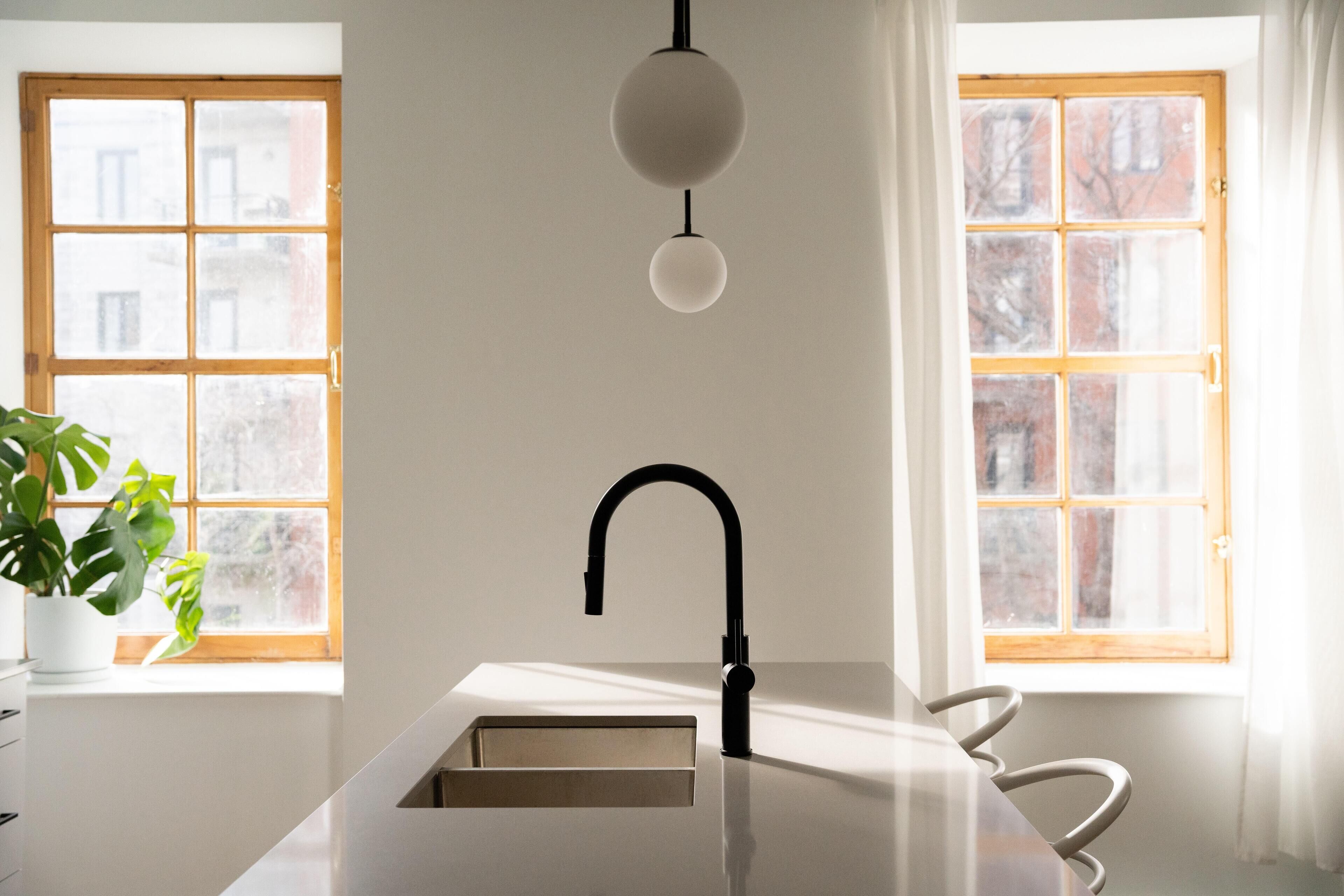 Une cuisine moderne avec un plan de travail blanc, un robinet noir et un luminaire suspendu. On y trouve deux fenêtres à cadre en bois et une plante sur le côté gauche.