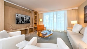 Une chambre d’hôtel moderne avec un canapé blanc, une table basse en bois, un téléviseur fixé au mur et une grande fenêtre ornée de rideaux légers.