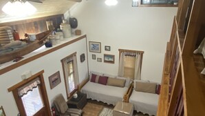 Amplia sala de estar con techo alto, vigas de madera y una cama tipo loft. Cuenta con ventiladores de techo, una mesa de comedor con sillas y un sofá.