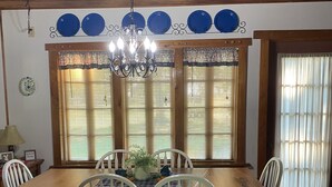 Un comedor con una mesa de madera y sillas blancas, rodeado de amplios ventanales con persianas.