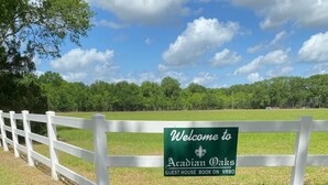 Un letrero da la bienvenida a los huéspedes en Acadian Oaks, con una valla blanca y follaje verde al fondo.