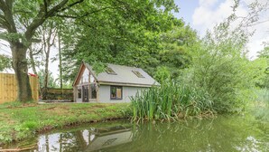 Une maison située au bord d’un étang paisible, avec une terrasse en bois et un mur de pierre en arrière-plan.