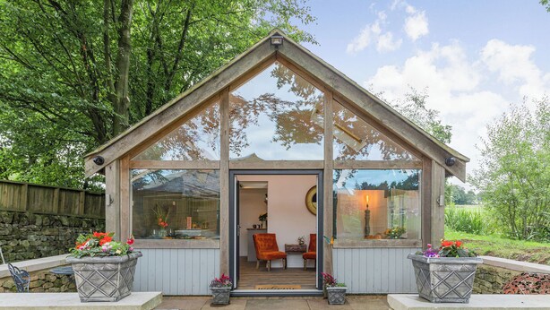Un belvédère à toit de verre, doté de fondations en pierre, de murs en bois et d’une porte donnant sur un intérieur comprenant une cheminée et des sièges.