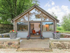 A glass-roofed gazebo with a stone foundation, wooden walls, and a door leading to an interior with a fireplace and seating. - 1 bedroom accommodation in Matlock (Matlock)