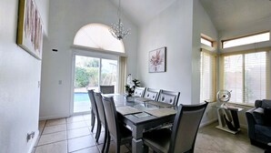 A dining area with a glass table, cushioned chairs, a chandelier, and a view of a swimming pool and greenery outside. - SantaFe Desert Oasis: Saltwater Pool, EV Charger, Near Coachella & Tennis Garden (Indio)