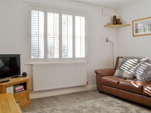 A living room with a television, a brown leather sofa, a window with blinds, a radiator, and a shelf with items. - 2 bedroom accommodation in Keswick (Keswick)
