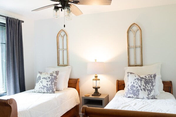 A bedroom with two beds, a ceiling fan, a nightstand with a lamp, and a window with curtains.