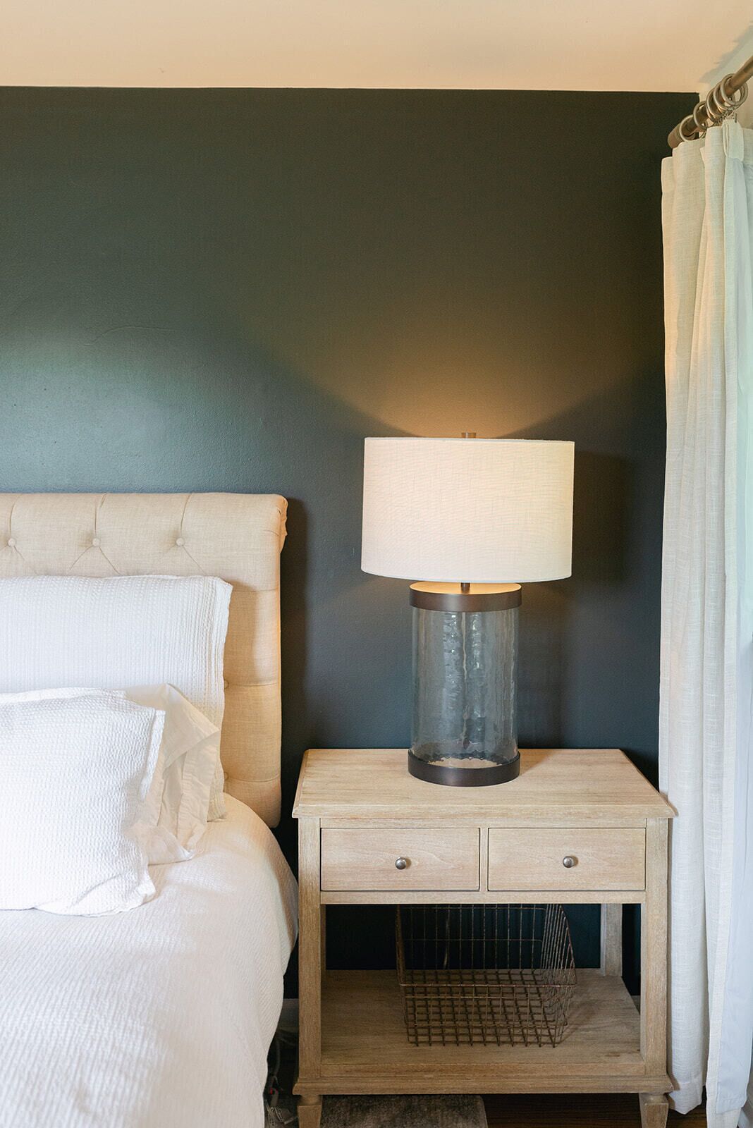 A bed with white linens, a bedside table with a lamp, and a curtain.