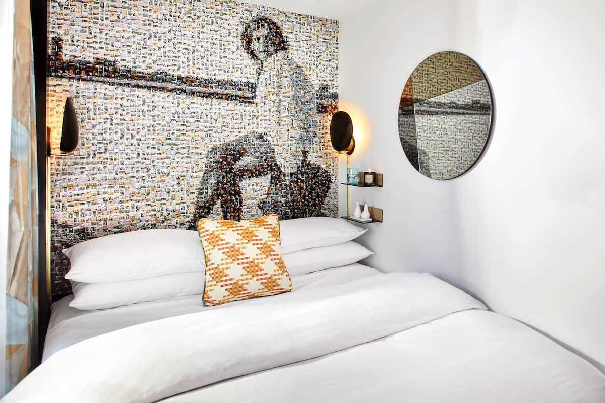 A hotel room with a bed, pillows, a wall art piece, a round mirror, and a bedside table with a lamp.