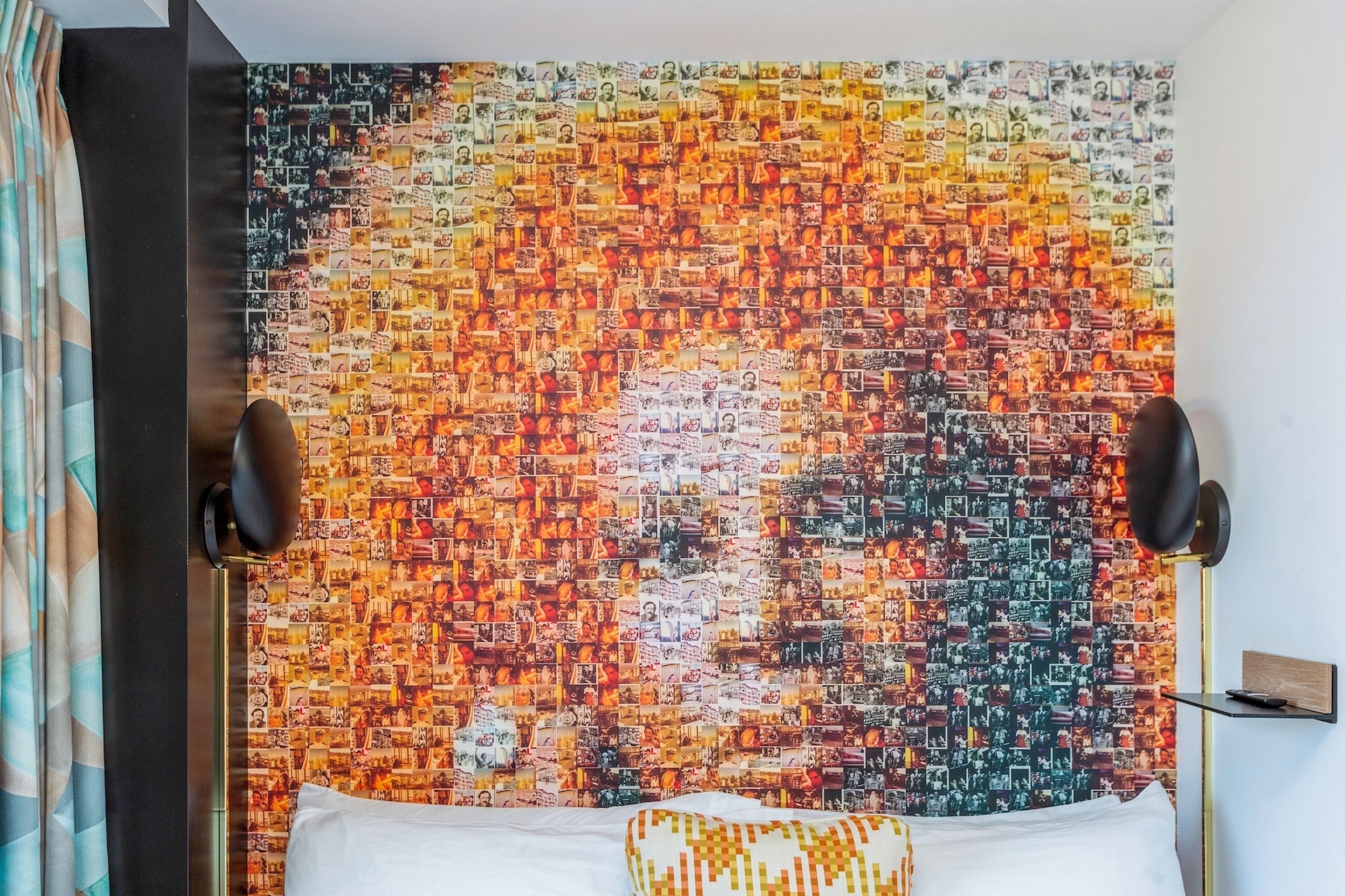 A room with a headboard covered in a collage of photos, two wall-mounted speakers, a bedside table with a lamp, and a patterned pillow.