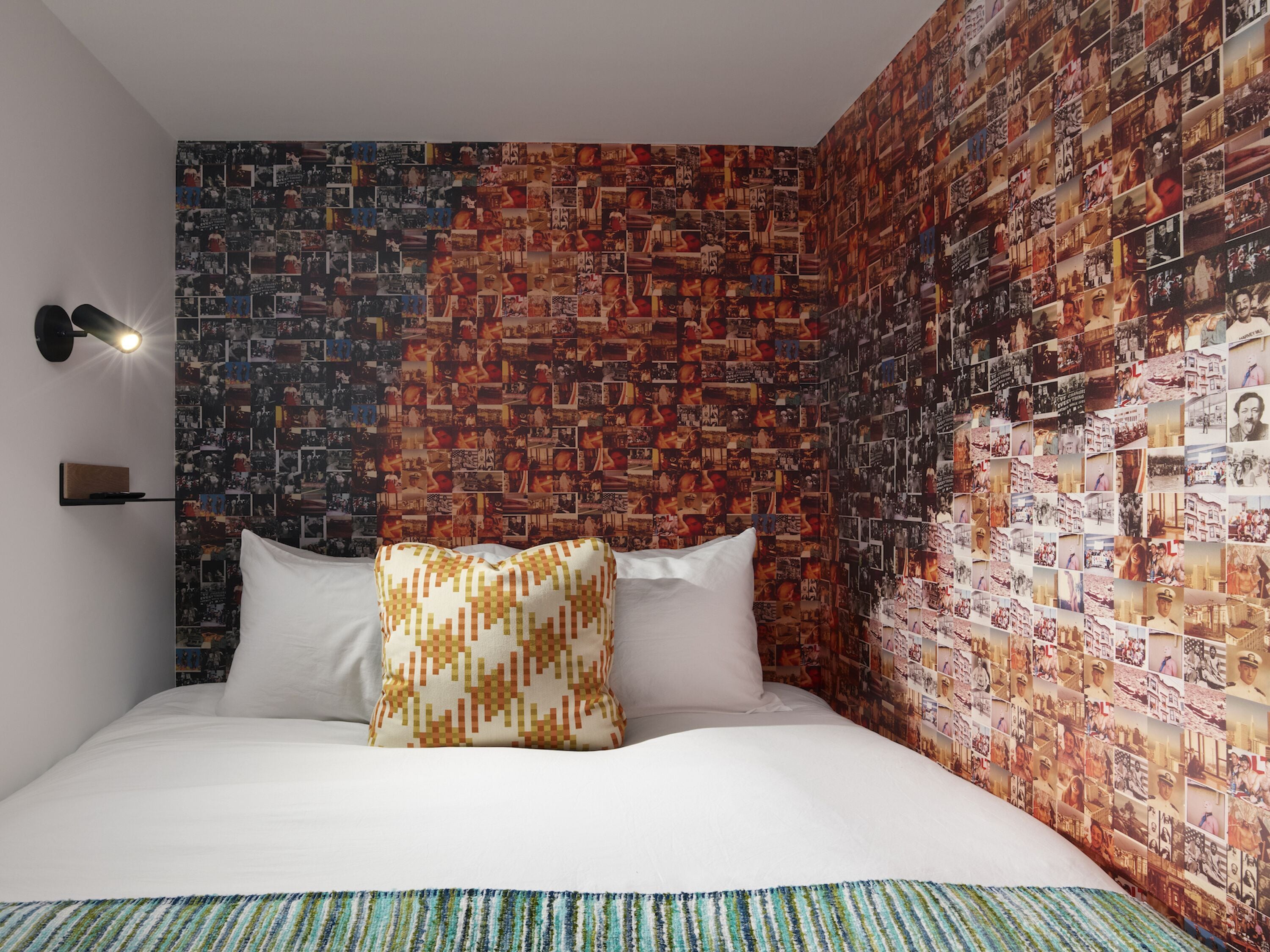 Une chambre à coucher avec un mur recouvert d’un collage de photographies, un lit avec du linge de lit blanc et une table de chevet avec une lampe.